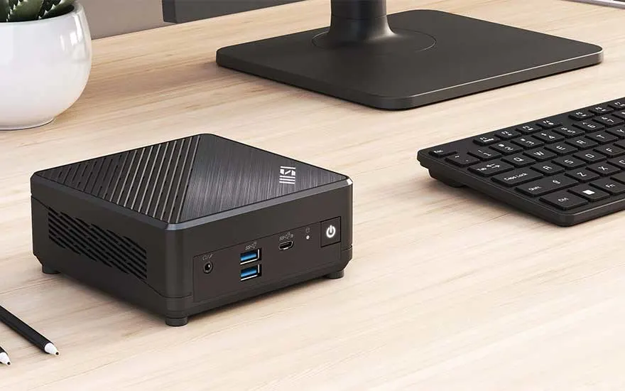 Zwarte mini-pc naast toetsenbord op een houten bureau, met een monitorvoet en een plant op de achtergrond.