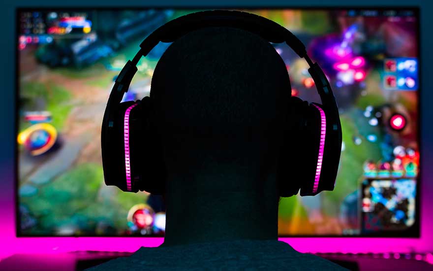 Een persoon met een gaming headset zit voor een scherm met een kleurrijk videospel. Roze verlichting is zichtbaar.