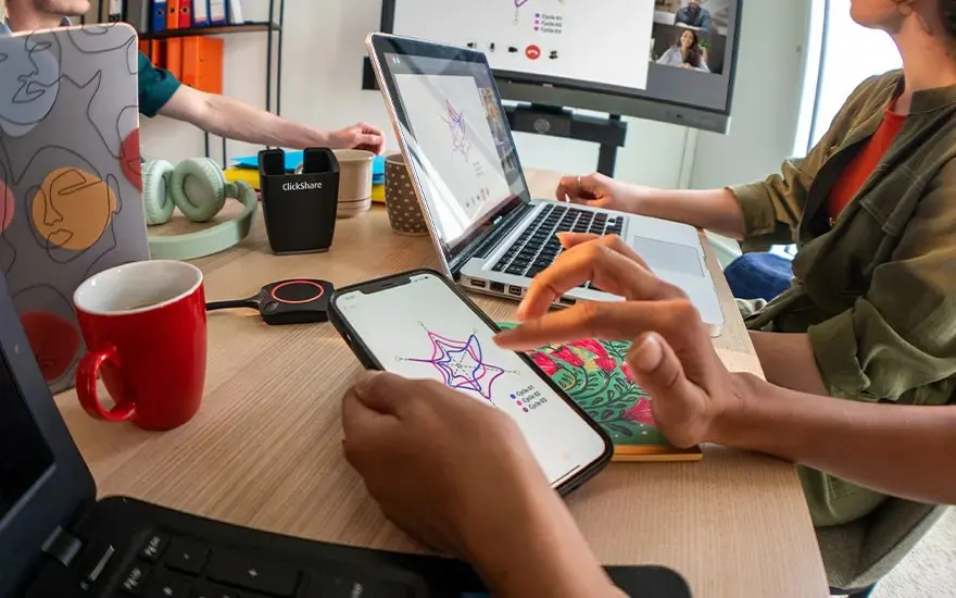 Several people collaborate with laptops, smartphones and a ClickShare button, with shared content shown on the screen.