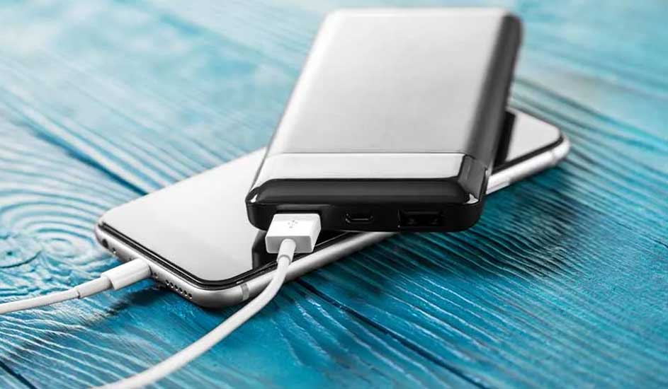 Un téléphone blanc se charge avec une batterie externe argentée sur une table en bois bleu. Un câble blanc relie les deux appareils.