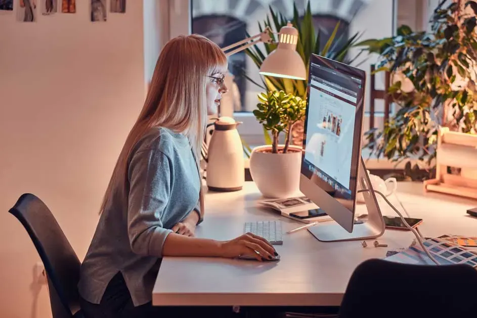 Une table blanche avec un ordinateur, clavier, lampe et plante en pot. Une fenêtre et des plantes sont visibles en arrière-plan.