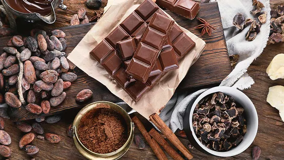 Een foto van chocolade, cacaobonen, cacaopoeder en kaneelstokjes op een houten tafel.