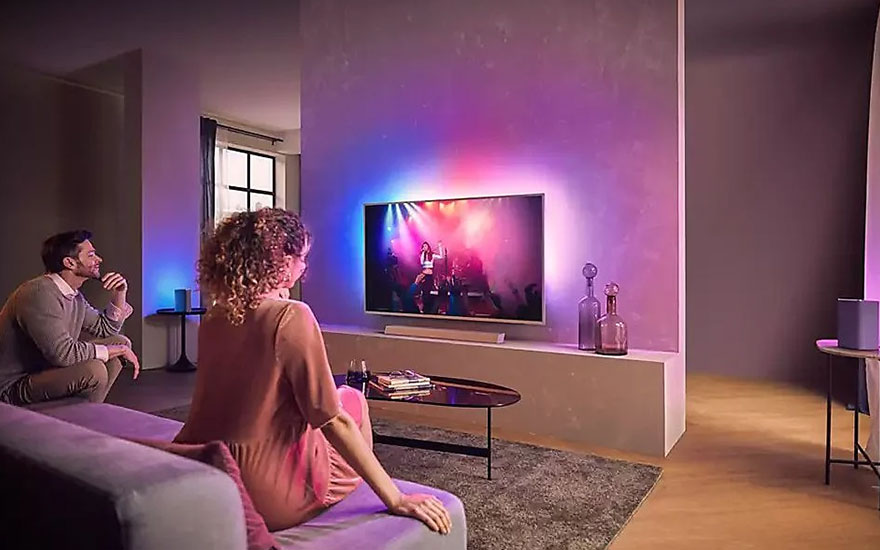 Woonkamer met tv aan de muur, paarse en blauwe achtergrondverlichting. Een bank, tafel en decoratieve flessen.