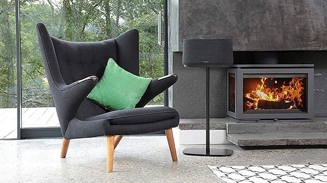 Fauteuil noir avec coussin vert près d'une fenêtre et d'un foyer allumé. Haut-parleur noir sur pied à côté.