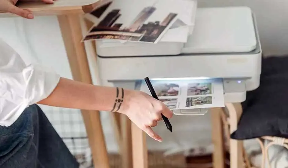 Een witte printer drukt foto's af. Een persoon houdt een zwarte pen vast. Op de achtergrond staat een houten standaard.