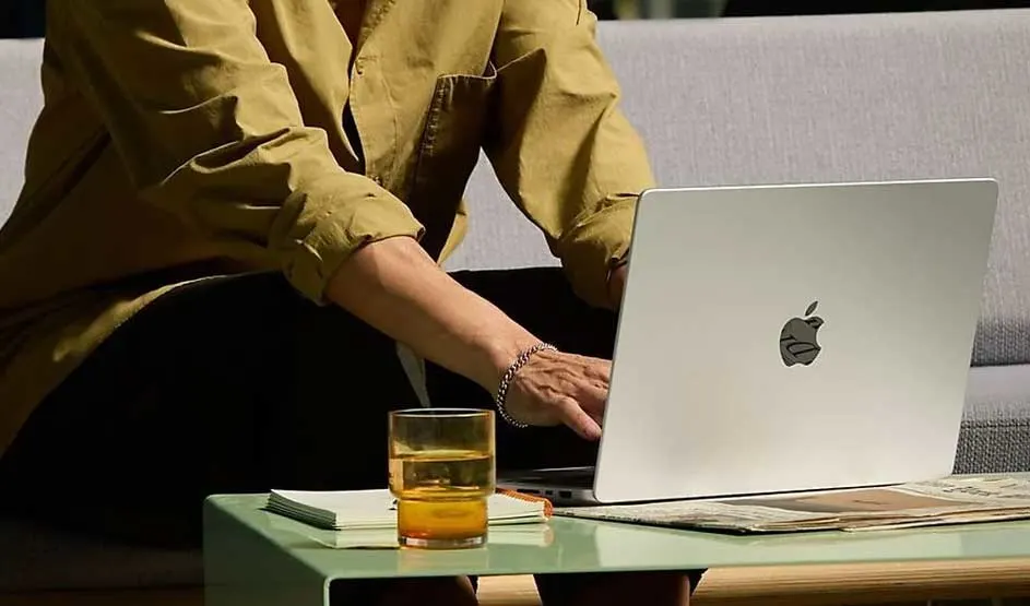 Personne en chemise jaune travaille dehors sur un MacBook Apple posé sur une table basse avec verre, carnet et magazine ; logo Apple visible.