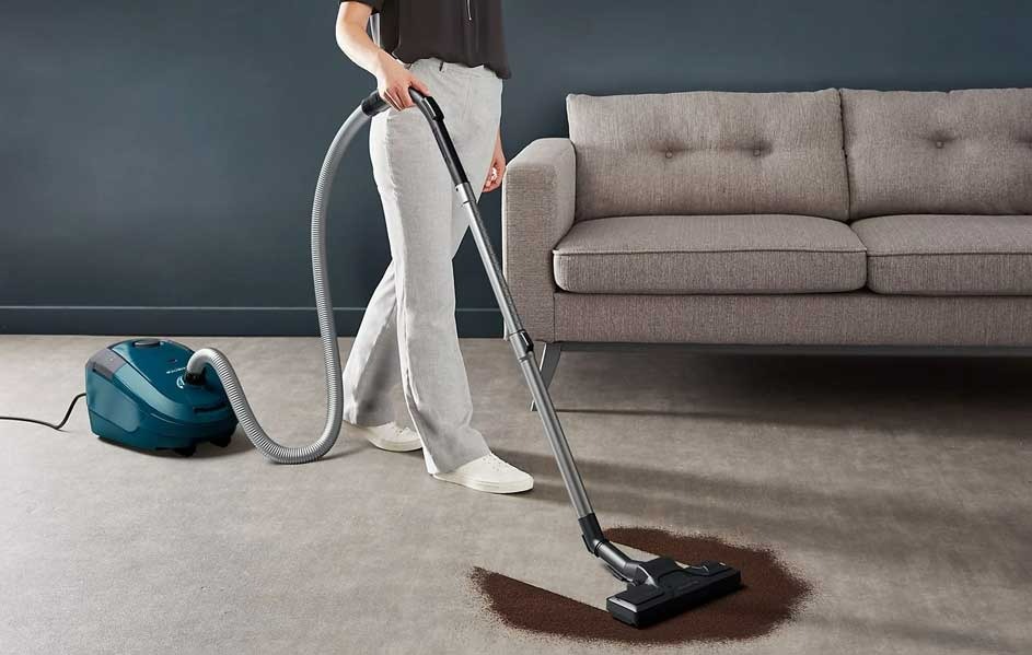 Een blauwe stofzuiger met een grijze slang reinigt een bruine vlek op een beige tapijt in een kamer met een grijze sofa.