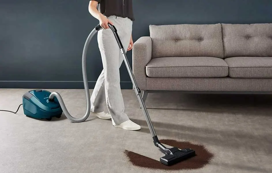 Een blauwe stofzuiger met een grijze slang reinigt een bruine vlek op een beige tapijt in een kamer met een grijze sofa.