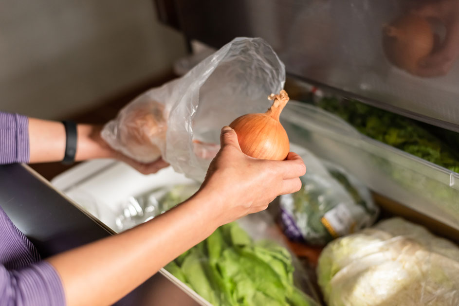 Une main tient un oignon brun près d'un sac plastique, au-dessus d'un tiroir de réfrigérateur rempli de légumes verts.