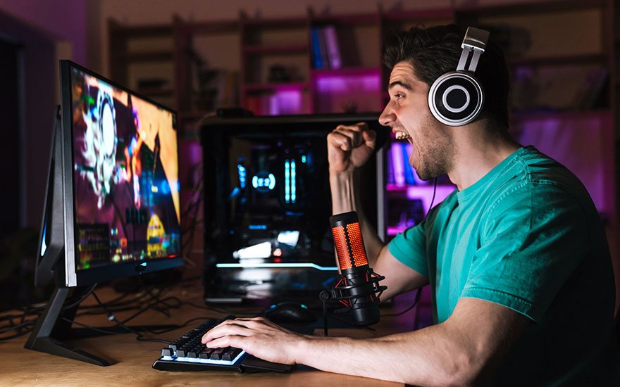 Un écran affiche un jeu vidéo, un clavier est posé sur une table en bois. Un microphone est visible.
