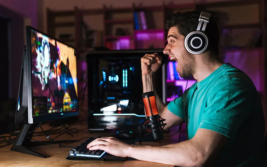 Un écran affiche un jeu vidéo, un clavier est posé sur une table en bois. Un microphone est visible.