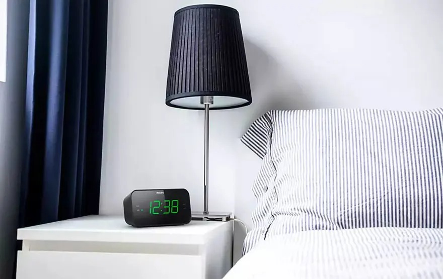 Een zwart Philips-wekkerradio op een wit nachtkastje naast een bed met gestreept beddengoed en een zwarte lamp. Merk: Philips.