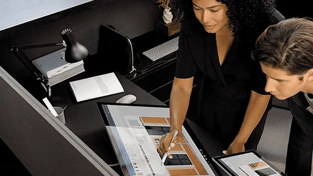 Deux personnes travaillent sur des écrans tactiles sur un bureau noir avec lampe, livres et clavier.