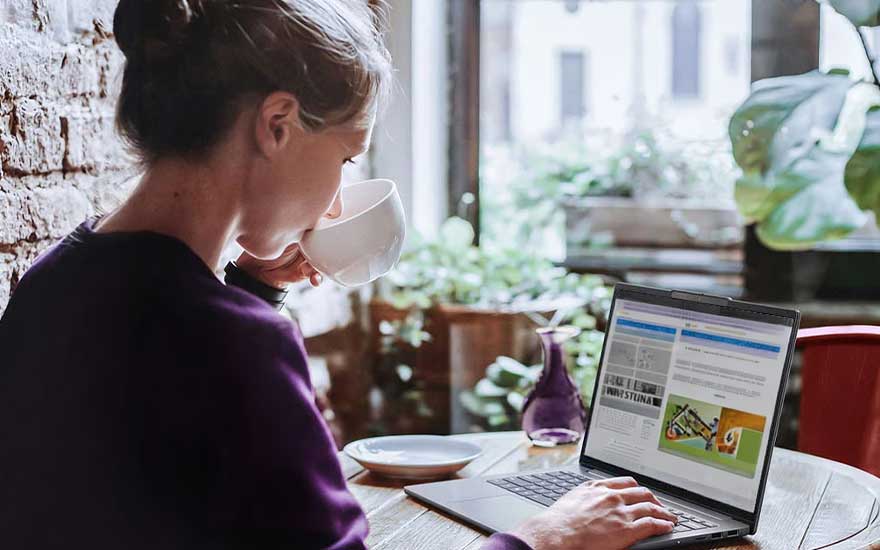 Een vrouw drinkt koffie aan tafel en bekijkt een webpagina op haar laptop, omringd door planten, servies en zacht daglicht.