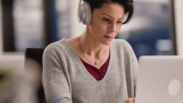 Une personne porte un casque gris et regarde un ordinateur portable. Elle porte un pull gris et un collier.