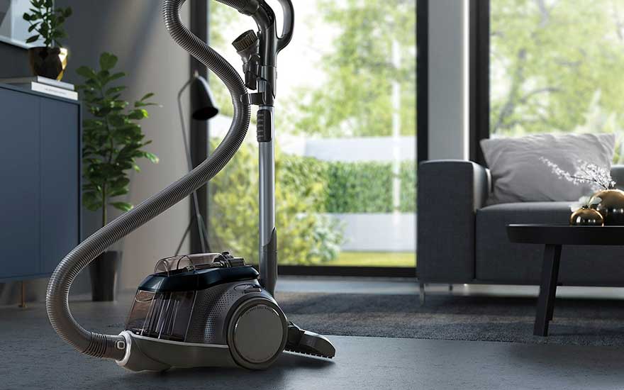 Un aspirateur gris et noir est posé sur un sol gris dans un salon moderne, près d'une fenêtre donnant sur un jardin.