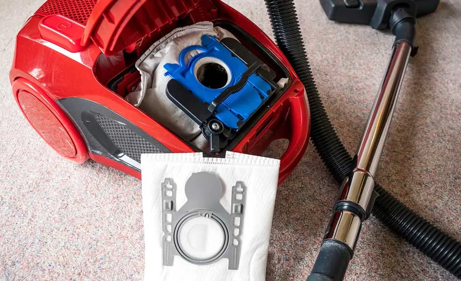 Een rode stofzuiger met open klep, een volle stofzak en een nieuwe witte stofzak op een beige tapijt.