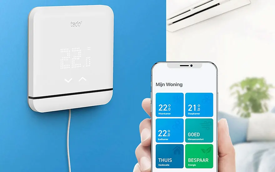 Witte thermostaat aan een blauwe muur, met een smartphone die een app toont met temperatuurregeling. Airco zichtbaar op achtergrond.