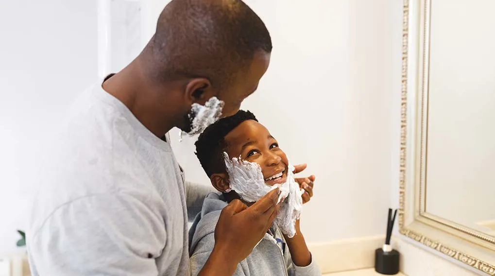 A man helps a child in the bathroom with shaving foam on their face, in a bright space with a mirror.