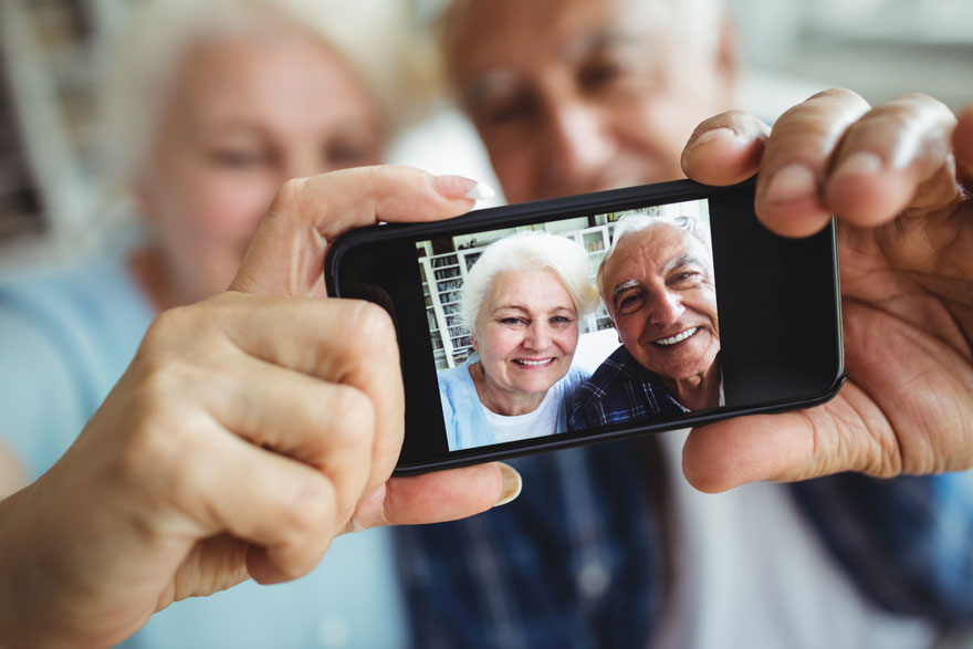 Un couple âgé regarde un téléphone montrant une photo d'eux-mêmes souriant devant un bâtiment blanc.