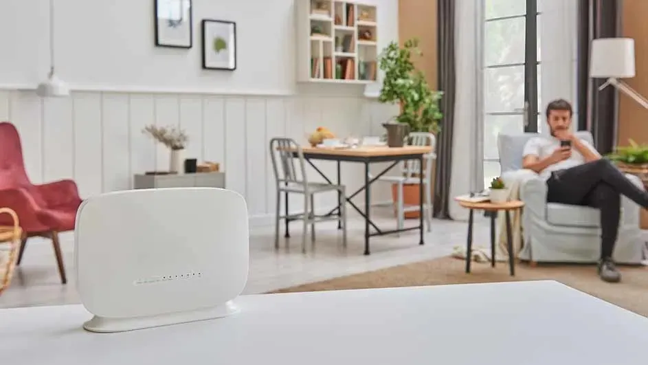 Un routeur blanc sur une table. En arrière-plan, une salle à manger, une fenêtre et un fauteuil.
