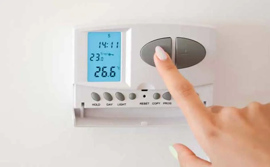 Un écran numérique affiche 14:11 et 26,6°C sur un thermostat blanc. Un doigt appuie sur un bouton gris sur le thermostat.