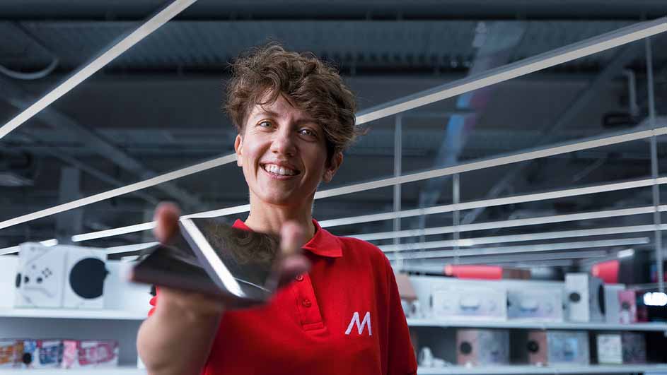 Een rode polo met een witte 'M' presenteert een zwarte tablet in een winkel met rekken vol elektronica en witte lampen.