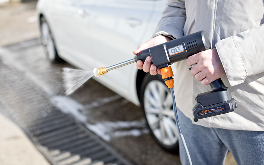 Des mains tiennent un nettoyeur haute pression portable noir dirigé vers une voiture blanche, le design compact de l'appareil étant bien visible.