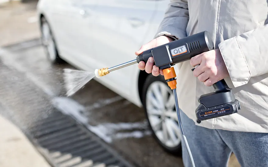 Des mains tiennent un nettoyeur haute pression portable noir dirigé vers une voiture blanche, le design compact de l'appareil étant bien visible.