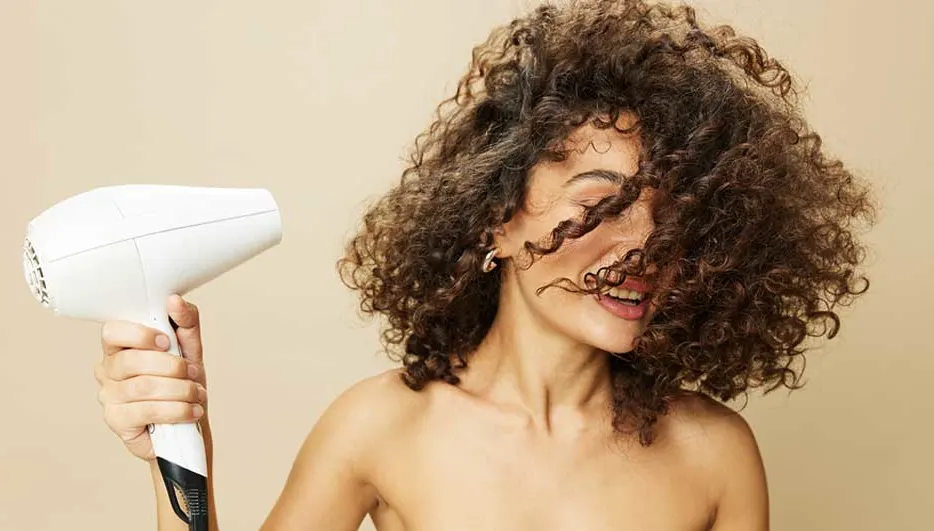 Une main tient un sèche-cheveux blanc sur un fond beige uni. Cheveux bouclés bruns.