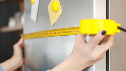 Une personne mesure un réfrigérateur argenté avec un ruban à mesurer jaune. Des notes sont fixées sur le réfrigérateur.