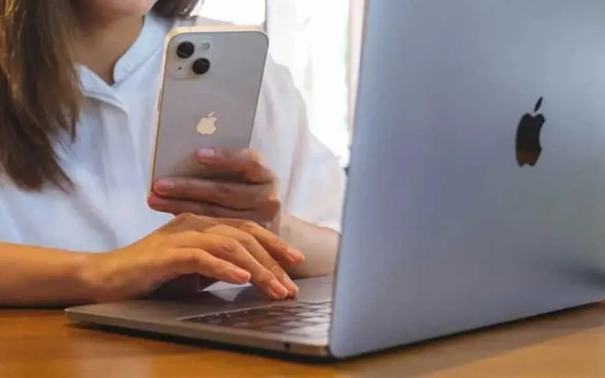 A person sits at a wooden table with an open MacBook, holding an iPhone upright in one hand while typing on the laptop keyboard with the other.