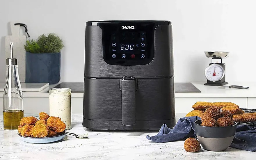 Een zwarte airfryer staat op een aanrecht, omringd door snacks, een fles olie, een pot saus en een weegschaal.