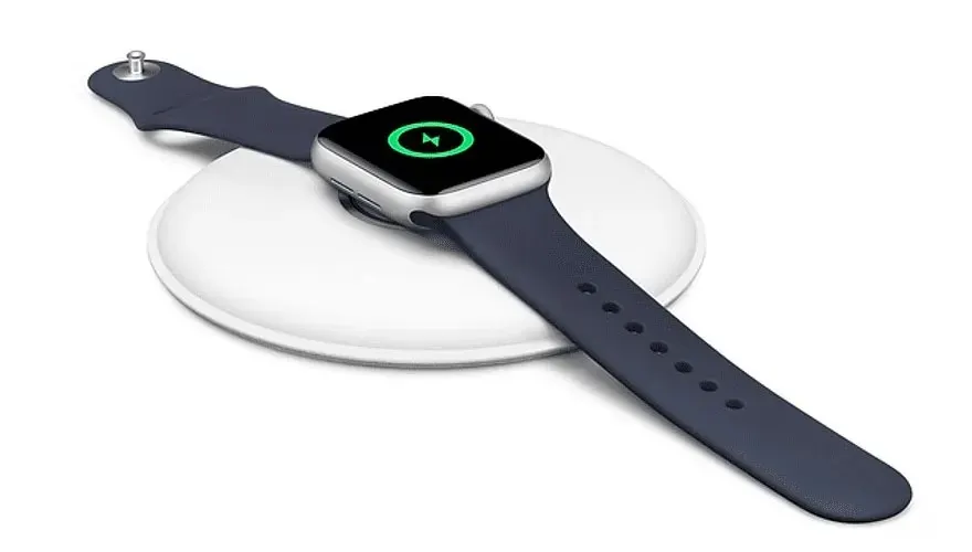 Een zilveren smartwatch met een blauw bandje ligt op een witte draadloze oplader. Het scherm toont een groen laadsymbool.