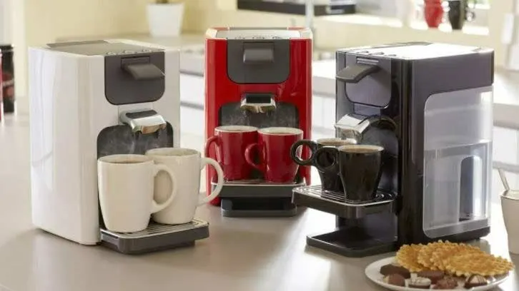 Trois machines à café (blanche, rouge, noire) avec des tasses assorties sur un comptoir. Des gaufrettes et des chocolats sont sur une assiette.