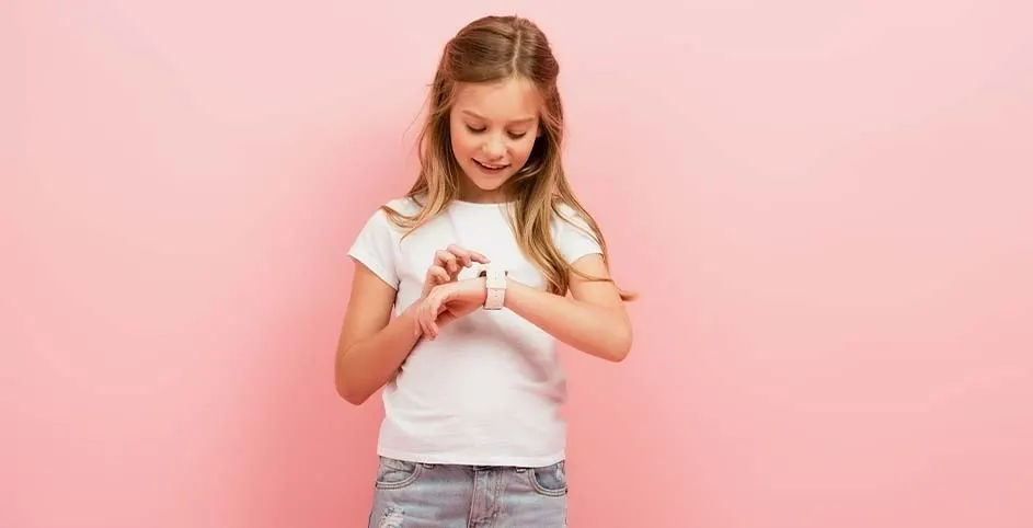 Een witte smartwatch op een pols tegen een roze achtergrond. De persoon draagt een wit t-shirt en een jeansbroek.