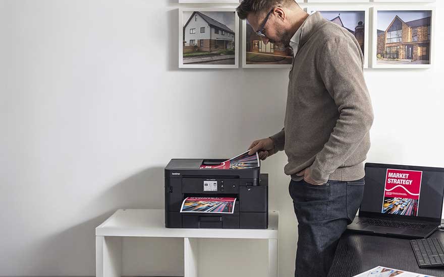 Een man gebruikt een printer terwijl kleurrijke documenten worden afgedrukt. Op de achtergrond hangen afbeeldingen van moderne woningen aan de muur.