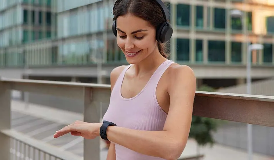 A woman in a pink sports top is wearing black Sony headphones and looking at her smartwatch on the street.