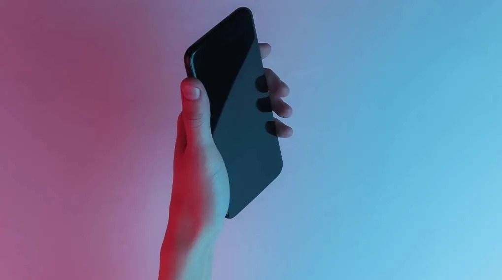 Une main tient un smartphone noir devant un fond rose et bleu.