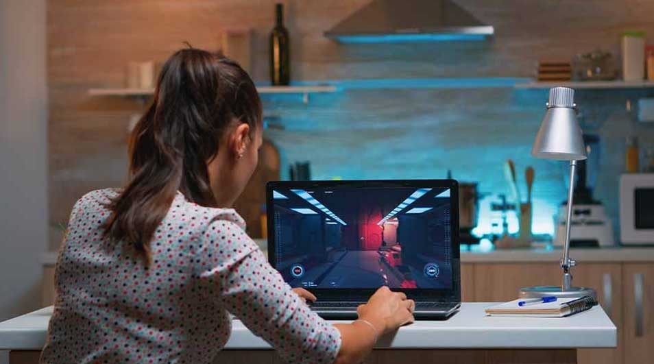 Laptop on a table displays a video game. A lamp and notebook sit beside it. The background shows a kitchen setting.