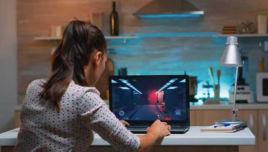 Laptop on a table displays a video game. A lamp and notebook sit beside it. The background shows a kitchen setting.