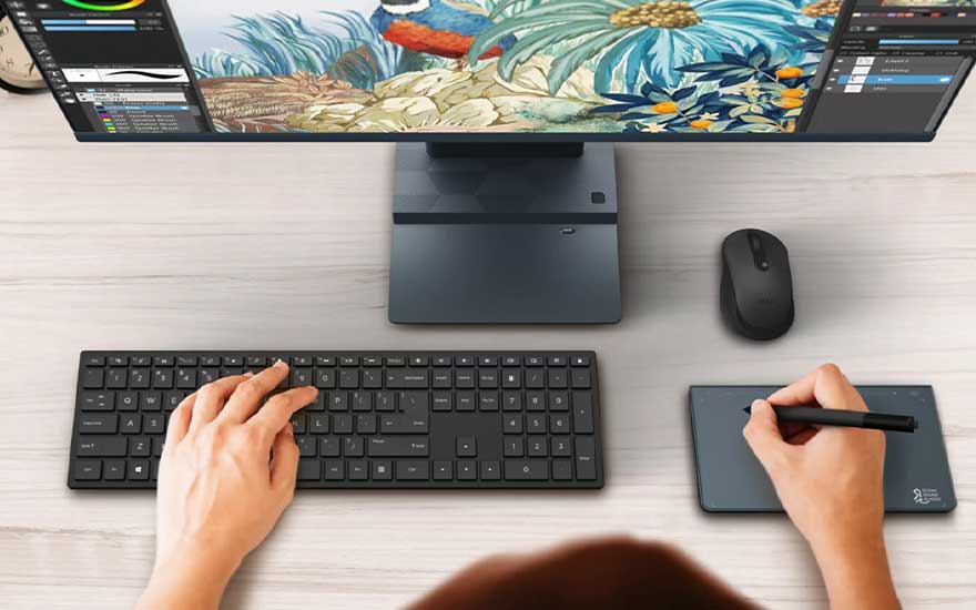 Un écran d'ordinateur affiche un motif floral coloré, avec un clavier, une souris et une tablette à stylet sur un bureau en bois blanc.