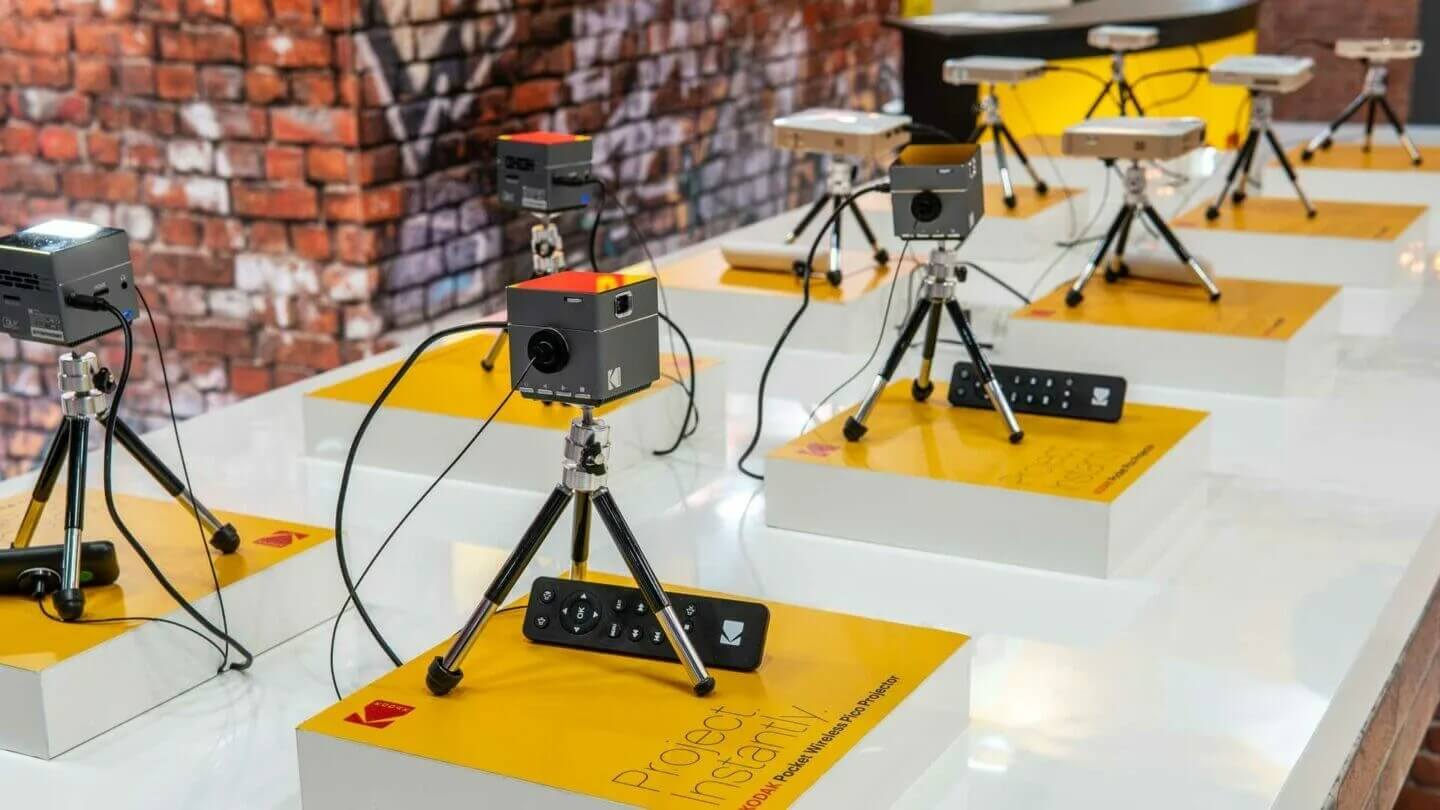 Rij met kleine projectoren op statieven, elk op een geel platform op een witte tafel tegen een bakstenen achtergrond.