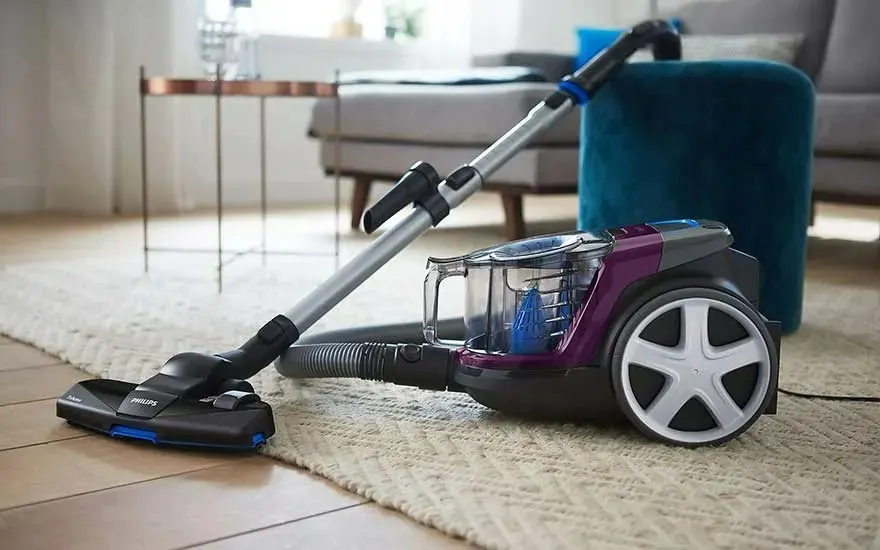 Un aspirateur traîneau Philips violet et noir sur un tapis clair, avec un tube gris et des meubles en arrière-plan.