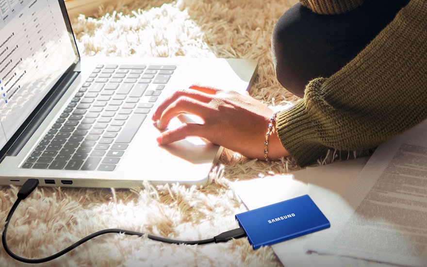 Laptop op een pluizig tapijt, verbonden met een blauwe Samsung SSD. Hand typt. Documenten zichtbaar.