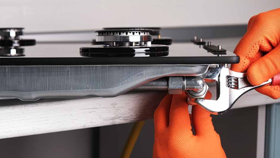 Close-up of a silver wrench tightening a gas pipe under a black stovetop with silver burners. Orange gloves are visible.