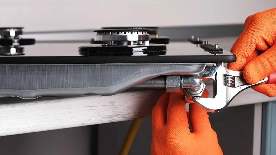 Close-up of a silver wrench tightening a gas pipe under a black stovetop with silver burners. Orange gloves are visible.