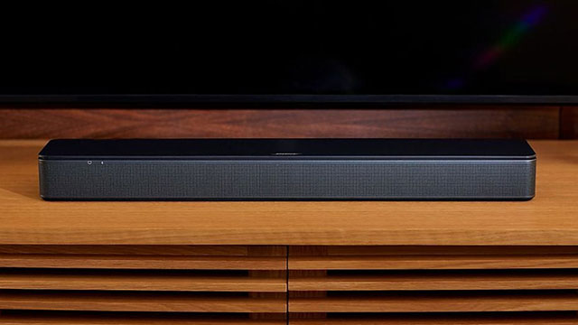 Een zwarte soundbar staat op een houten meubel voor een donker scherm.