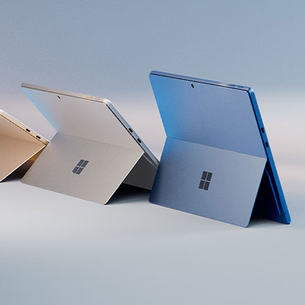 Drie Microsoft Surface-tablets in goud, zilver en blauw staan open op een lichtblauwe ondergrond. Elk scherm toont het Windows-logo.