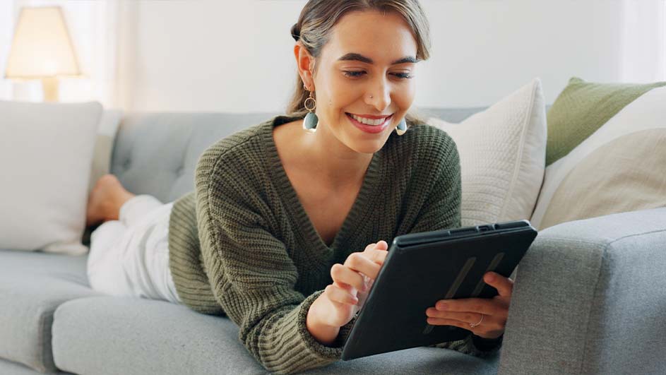 Une tablette noire est utilisée sur un canapé gris avec des coussins blancs et verts, sous une lampe.